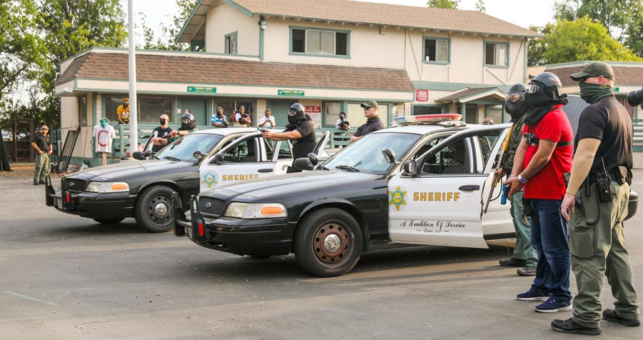 Civilians are outside infront of a Sheriff vehicle with it's doors open. Civilians are wearing protective head gear holding training pistols. Training deputies are on the side of them providing instruction.