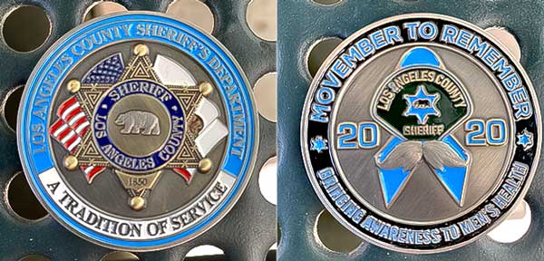 Front side of the coin has the sheriff's star siting over the American flag and the California State flag, the words "A Tradition of Service" along the bottom. The back of the coin has the LASD Cancer Awareness patch with a moustache underneith it, The words "Movember to Remember, Bringing Awareness to Men's Health Runs around the coin.