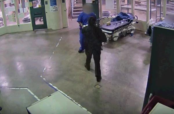 Still from security camera showing a Sheriff's Custody Assistant in uniform handing an inmate an item just before she is attacked. The inmate is dressed in a blue jump suit and is standing infront of a gurney. The room has a wide concrete floor and is surrounded by glass walls and a door is behind them.