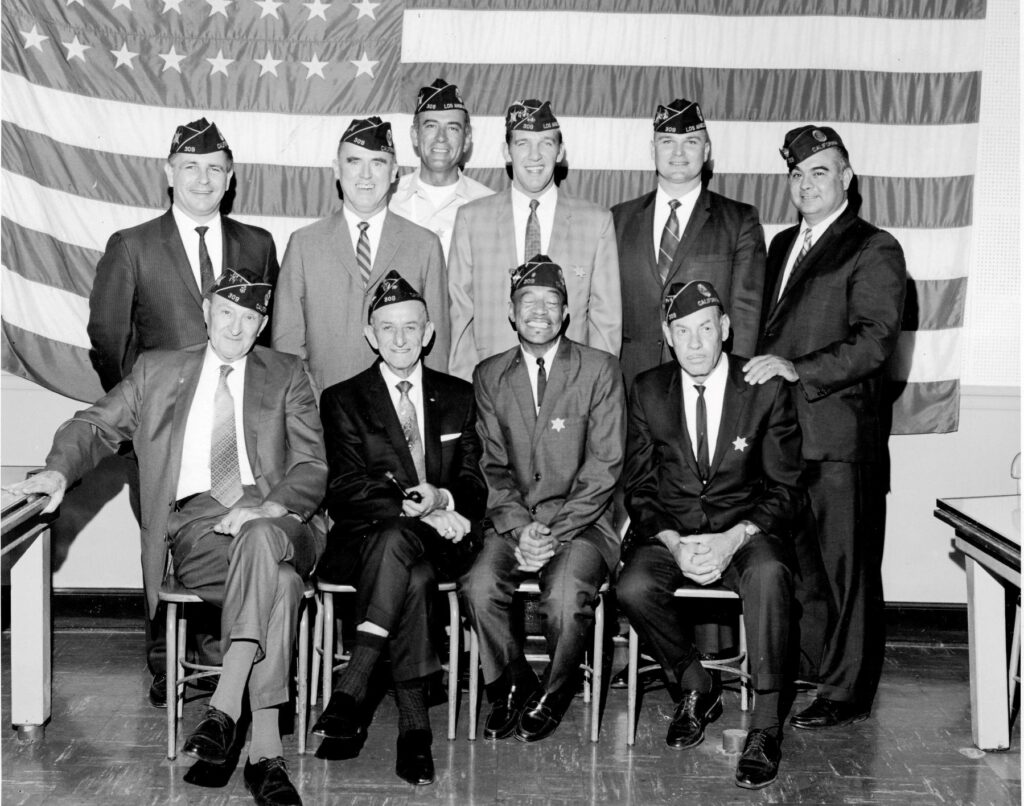 vintage black and white photo of 10 men in stuits with legion cover hats on. Posing shoulder to shoulder infront of a wall sized american flag.