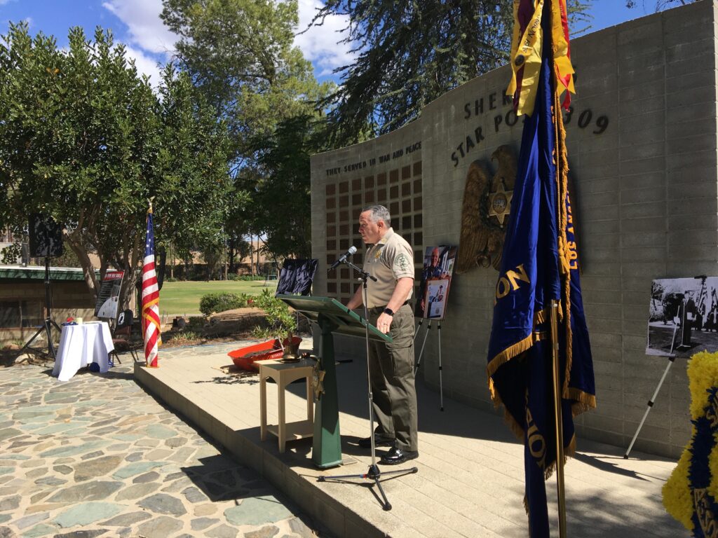 Image of Sheriff Alex Villanueva giving speach infront of the Star Post Memorial wall at a podium between the american flag and the American Legion flag.