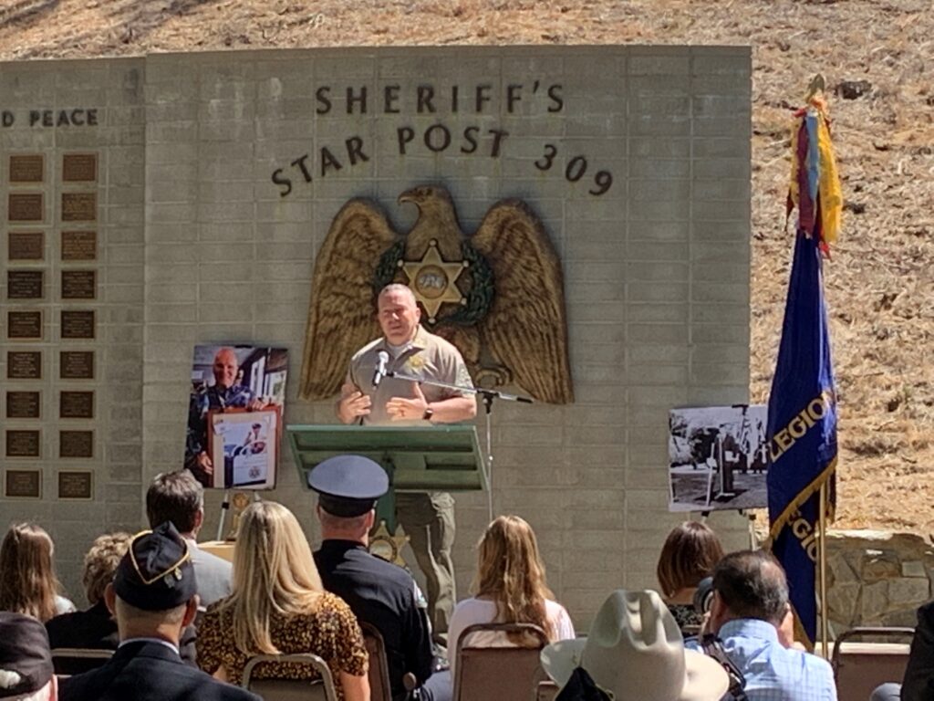 Sheriff Alex Villanueva standing infront of the Star Post 309 Memorial wal giving a speech infront of a crowd.