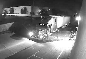 image of black and white security camera footage of a large Semi Rig driving through a ro Iron Gate in a back alley of a business. The semi has its lights on and is pulling a long 45 foot white trailer.