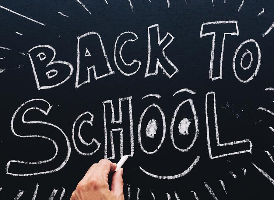 image of a hand writing on a chalk board, the board reads "BACK TO SCHOOL" and has a smile under the two o in the word "School".