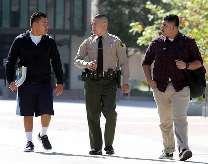 deputy sheriff is walking with two hispanic men down the street towards a camera. They are having a friendly conversation. The Deputy is wearing a tan longsleeve shirt with a black tie and green pants. there are two men on either side of the deputy. They are holding books and a backpack.