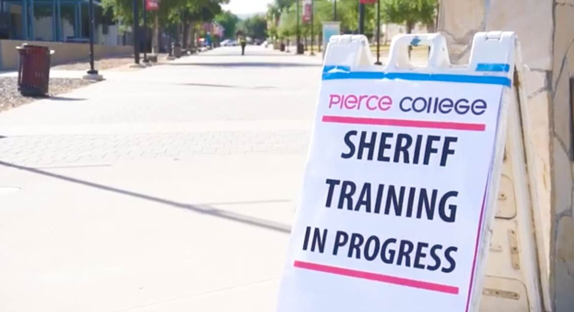 Image of an A-frame sign with big black letters that read "Sheriff training in progress". The sign in the lower left side of the frame. The sign is infront of a college walk path.
