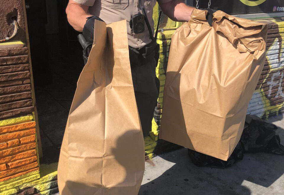 deputy is walking out of a business door holding two paper bags full of confiscated drugs.