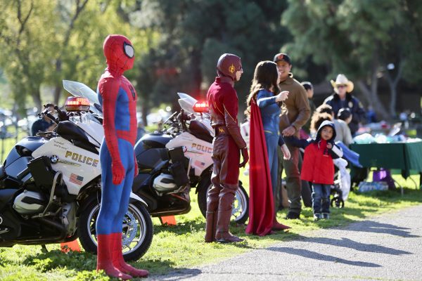 outdoor picture of Sheriff's motorcycles parked along a race path. People dresses as Spiderman, Flash and Supergirl are standing facing the racers.