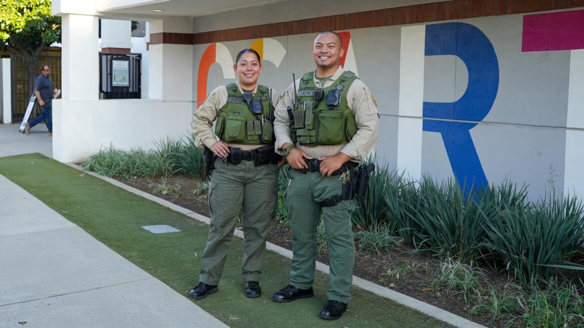 Two deputies are standing side by side, A male and female deptuy are wearing Green Vests and green pants and Tan Long sleeve shirts. They are standing infront of a building that has large multi colored letters on the wall.