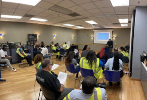 Over 20 people are sitting in a class room with a wooden floor. Looking towards a person at a podium. The person is standing infront of a projection screen.