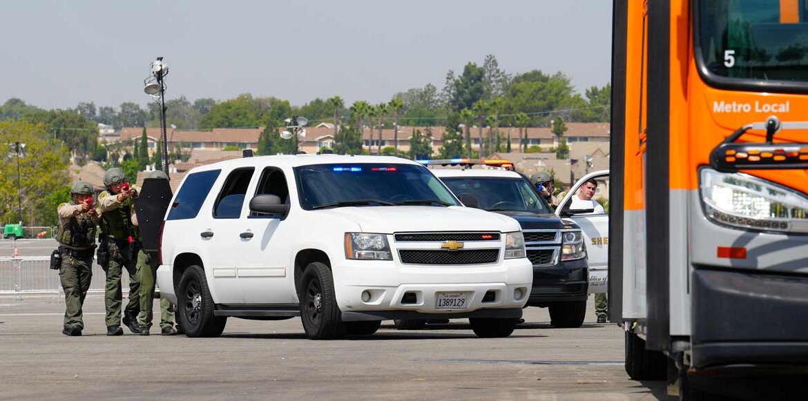 Three deputies in safety gear pointing training weapons from behind a white SUV at a bus parked in a parking lot training exercise.