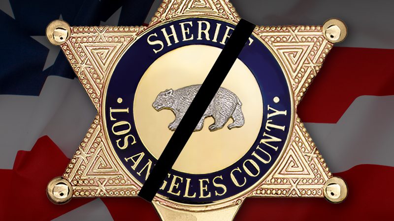 Los Angeles County Sheriff's Badge, Gold, six point badge. Sheriff, Los Angeles County is written in the center blue line of the badge, and a bear engraving is in the center, the bear is on all fours and walking left. There is a black band on the badge going right to left, diagnally, The badge sits on an image of the american flag.