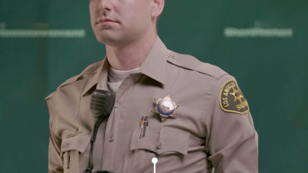 Class A Patrol Uniform: deputy with sandy blond hair is posing. he is wearing a tan wool longsleeve button down shirt. there is a gold LA County Sheriff's badge over his left pocket. There is a black rectangluar Body Camera clipped to his shirt in the middle. He has a walkie Talkie clipped to his lapel.