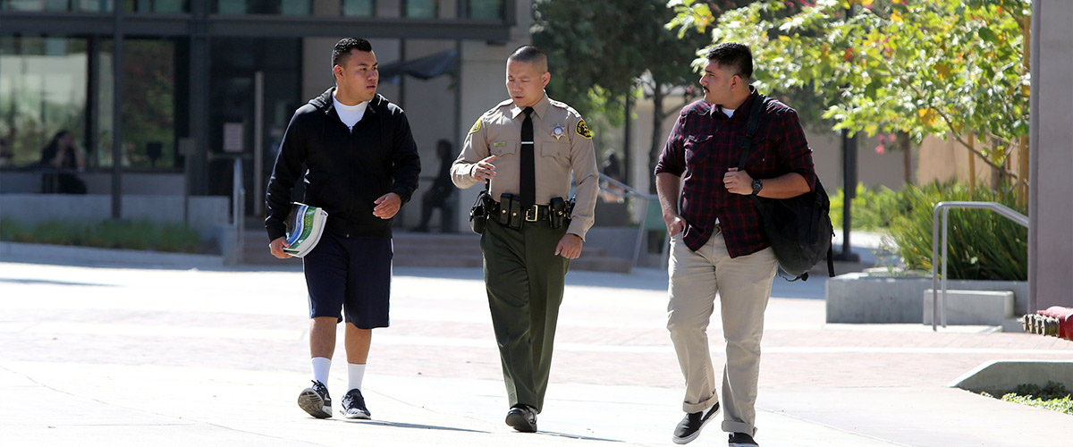 Image of a deputy dressed in green slacks, tan long sleeve shirt and black tie. walking with two hispanic students. The students are on either side of the deputy carrying books and book back slung across one student's sholder.