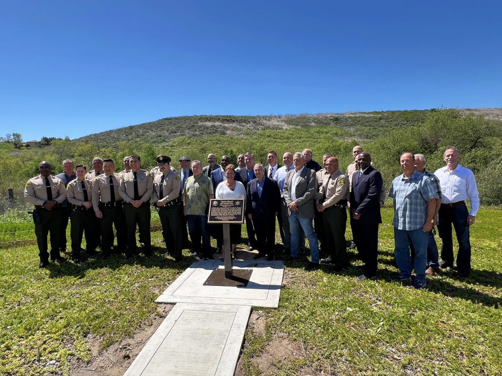 image of memorial site, city leaders and Sheriff's department personnel are posed behind the plaque in a field. 
