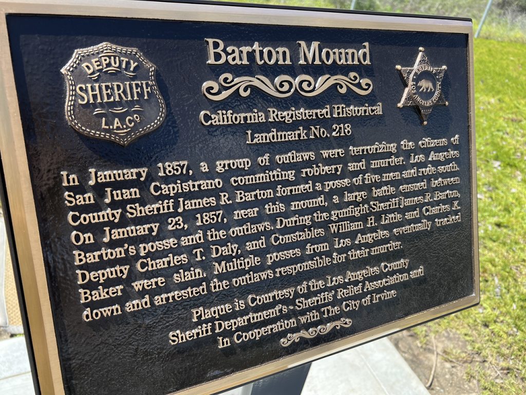 image of memorial plaque. Black background and gold letters. relief of old sheriffs badge on left and modern badge on right. Barton mound - California Registered Historical Landmark No. 218. In January 1857, a group of outlaws were terrorizing the citizens of San Juan Capistrano committing robbery and murder.