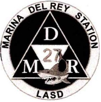 Circular logo with a triangle in the center. Marina Del Rey Station is in the outer most ring with LASD along the bottom. Inside the triangle, Letters D M R are in each Corner of Triangle. Number 27 for the station number is in another triangle in the center. There is a illustration of a shark inside the bottom of the circular logo.